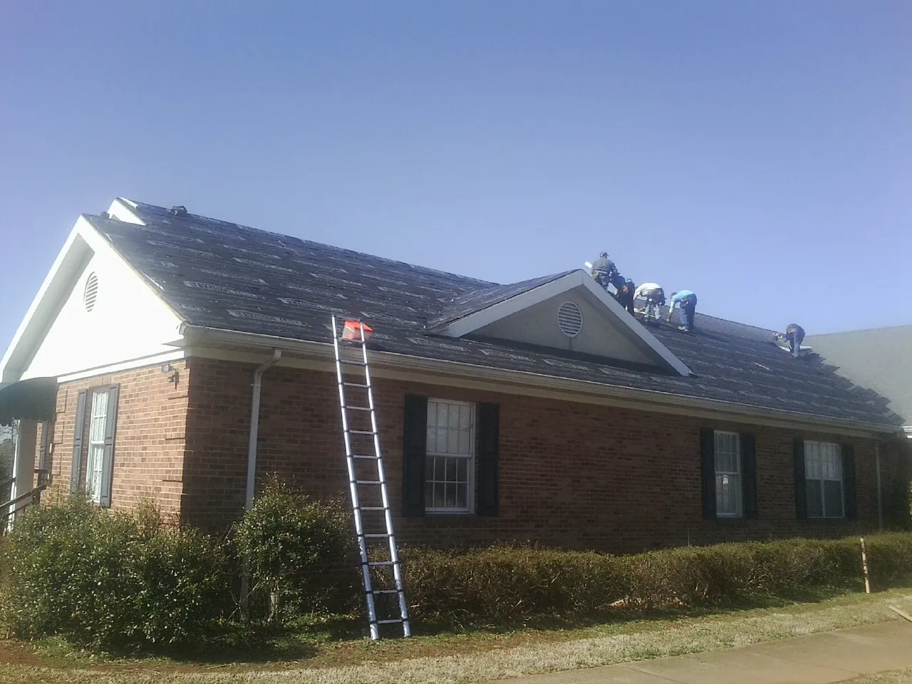 Crew tearing shingles off a brick ranch roof with a ladder set — Emergency Roof Repair work by our crew in Spangler