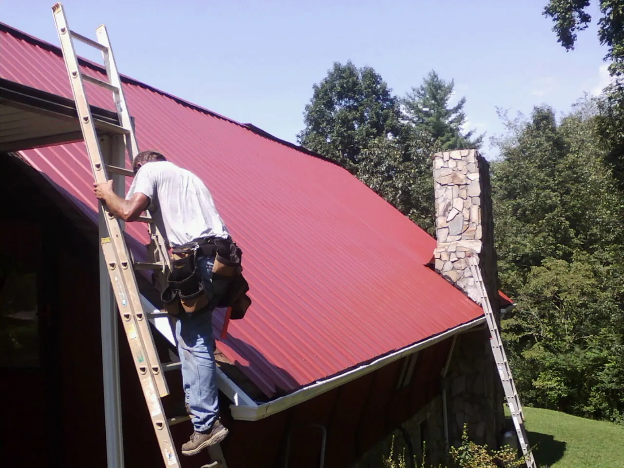 Roofer on a ladder beside a red metal roof and stone chimney — Soffit & Fascia Repair by Roofing Brothers in Spangler